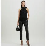 A stylish woman stands confidently in a minimalist studio. She wears a black pleated halter Women's Tops with a bow detail at the neck, paired with sleek black leather pants. Her accessories include a small black handbag with a gold clasp and strappy black high-heeled sandals. The background is a soft, neutral color, highlighting her chic outfit.