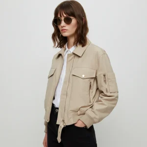 A young woman stands confidently, wearing a light beige bomber jacket with multiple pockets and a zippered front. She has shoulder-length, wavy brown hair and is accessorized with round, dark sunglasses. Underneath the jacket, she wears a crisp white shirt, and her outfit completes with high-waisted black pants. The background is minimalistic and neutral, emphasizing her stylish appearance.